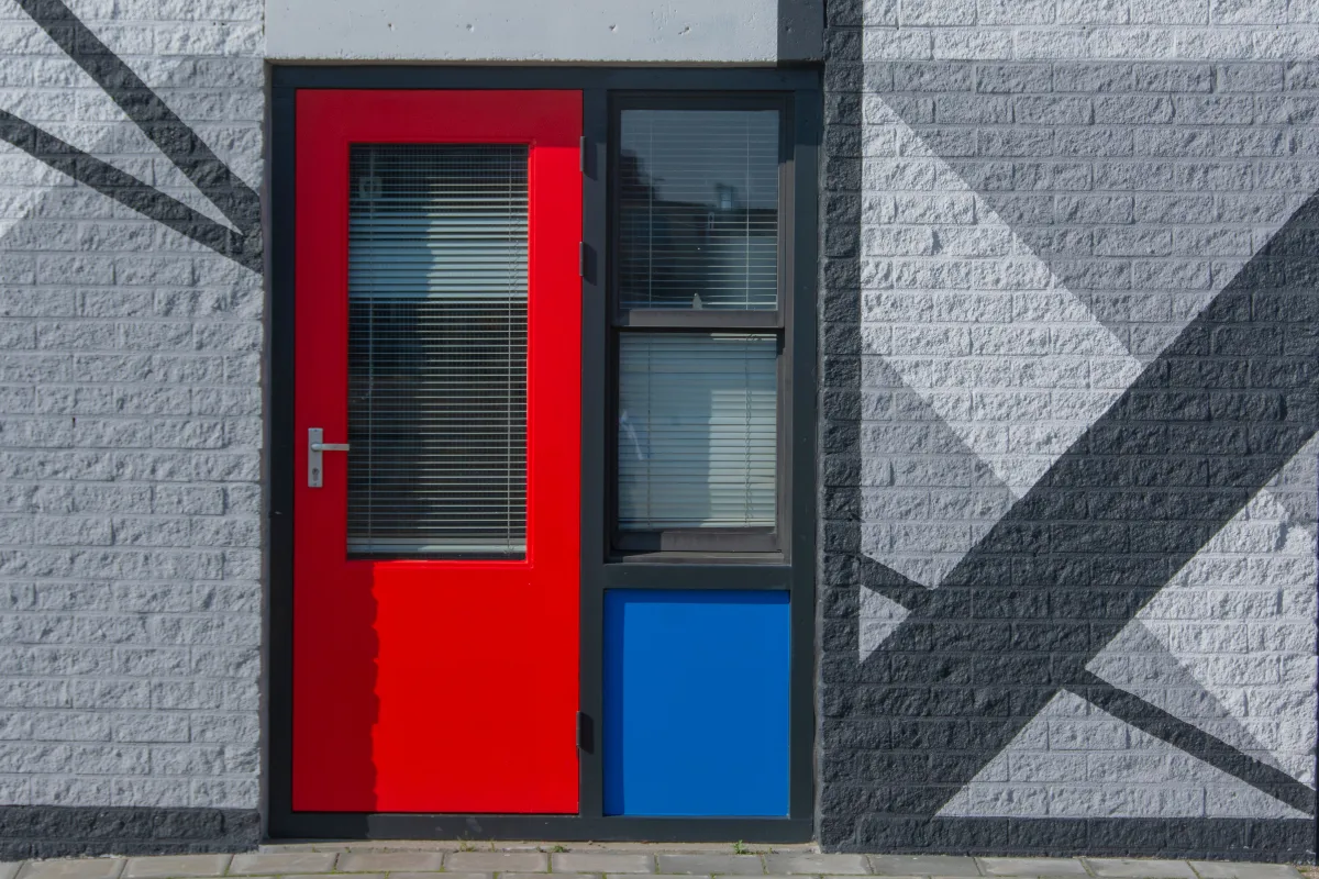 Hochwertige Lackierarbeiten: Eine leuchtend rot lackierte Außentür mit blauem Akzentpaneel vor einer geometrisch gestrichenen Backsteinwand.